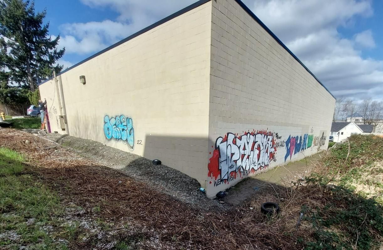 Graffiti on beige building exterior; brush, gravel, and shrubbery surround it. Blue sky overhead.