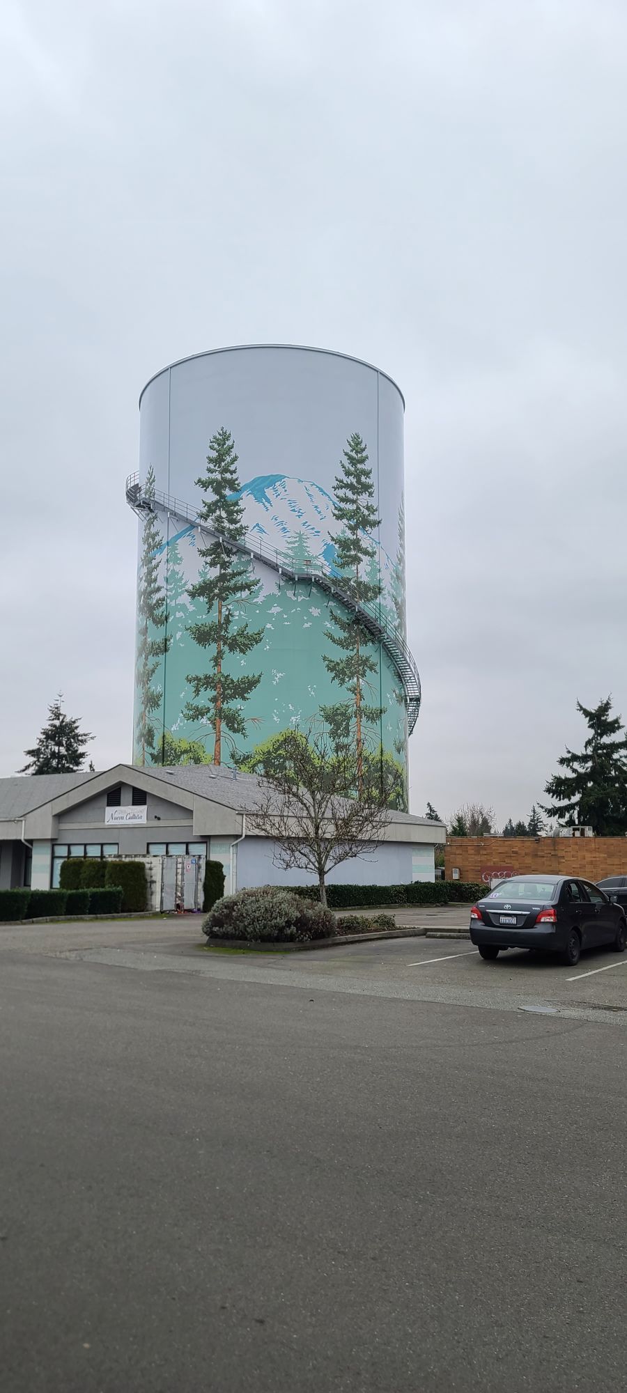 Water tower with a painted forest scene, next to a small building and parked cars. Overcast sky.