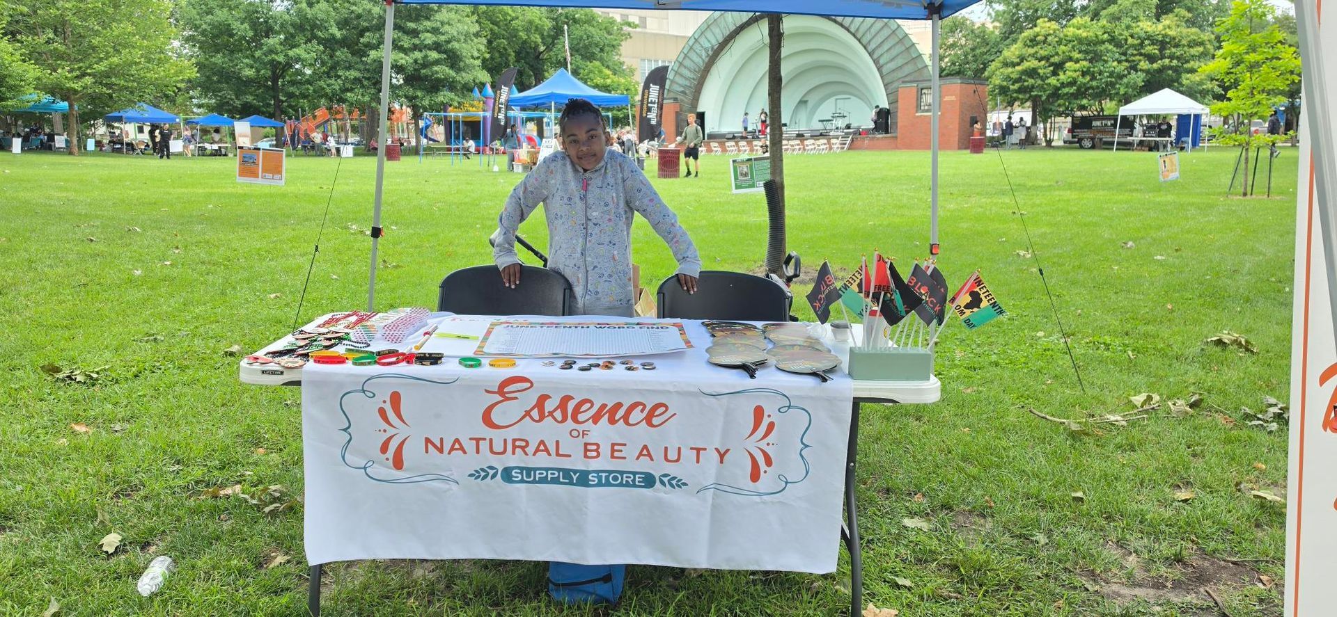 Vendor at an outdoor park booth with a white tablecloth and flowers, under a canopy on a grassy field