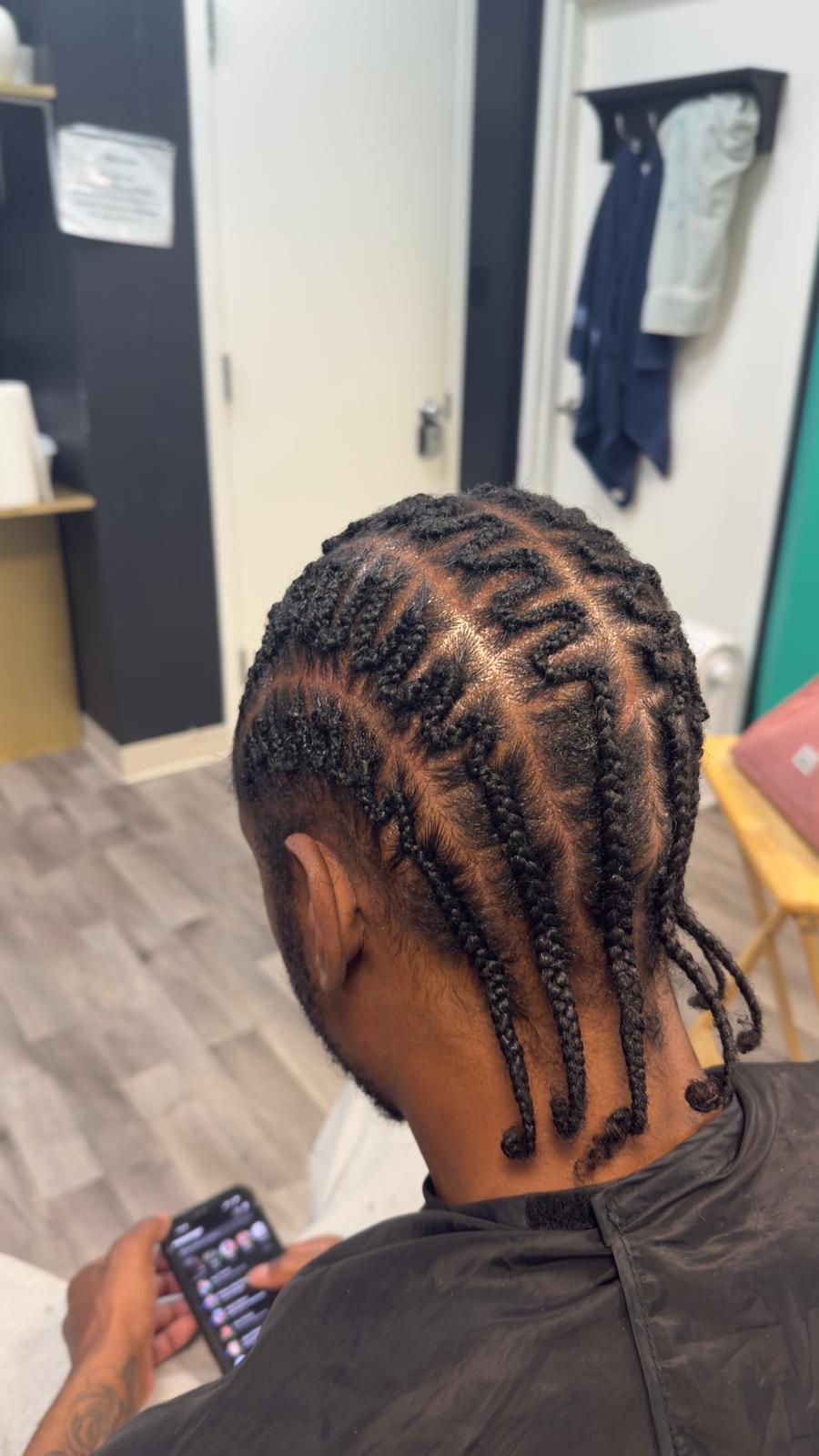 Person with braided hair looking at a phone in a room with open doors and hanging clothes.