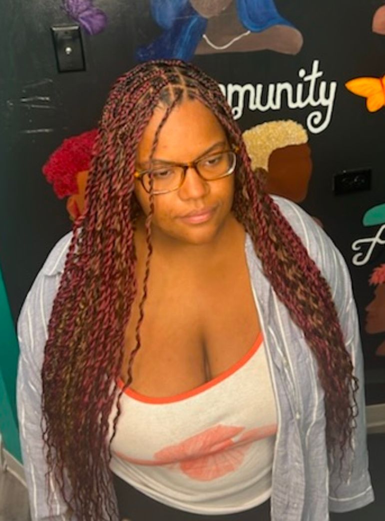 Woman with long red braids and glasses, wearing a white tank top, standing before a dark wall with colorful decor.