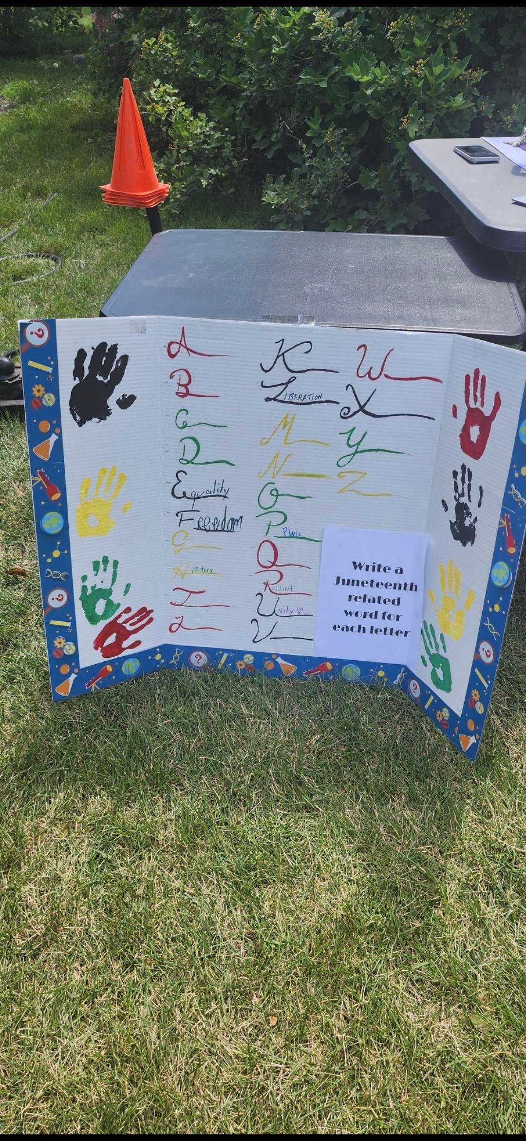 Colorful handmade poster on grass with drawings and handwritten notes beside a sidewalk and orange cone