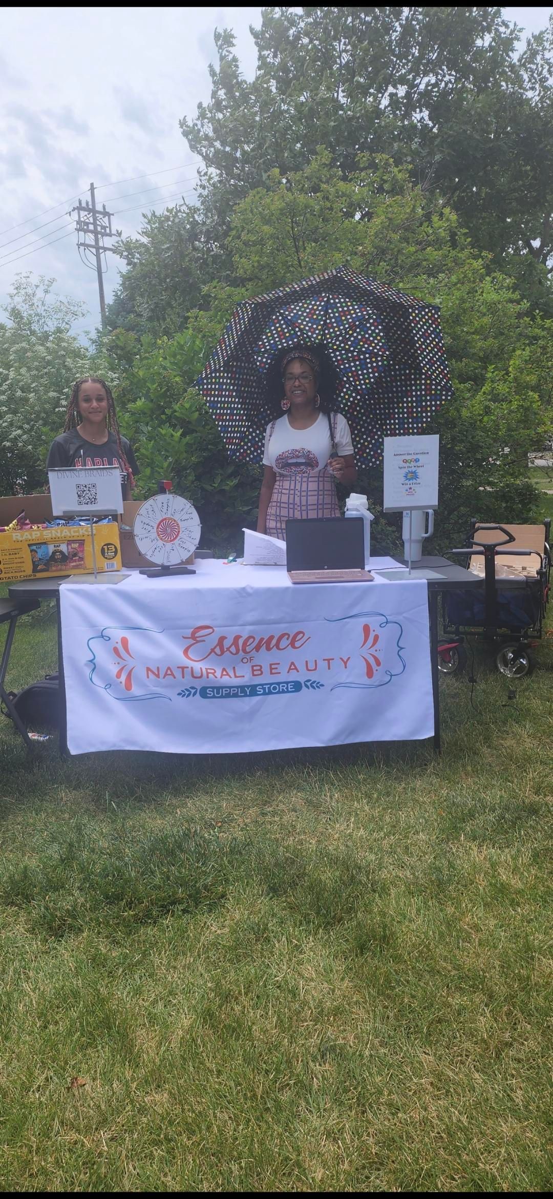 Outdoor vendor booth with a black-and-white umbrella, table displays, and a white banner in a grassy area.