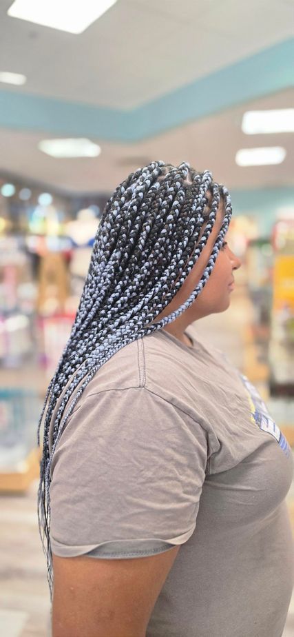 Profile of a person with long braided hair in a gray shirt, standing indoors with blurred store shelves in the background