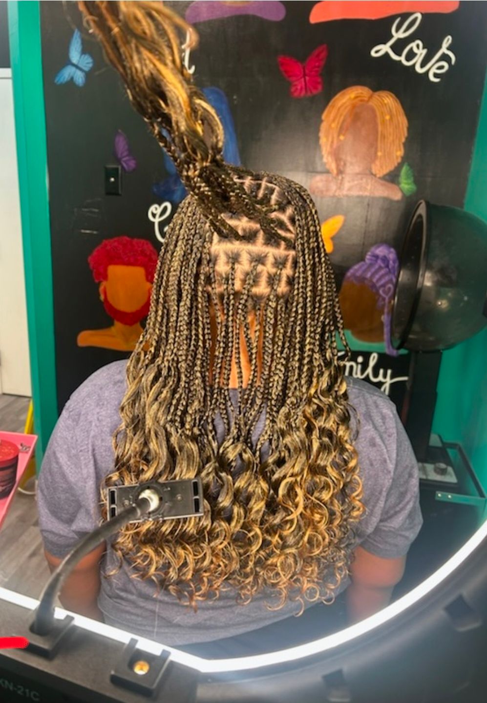 Person with long braided hair seated in a salon chair, seen from behind, with a mirror and neon sign ahead