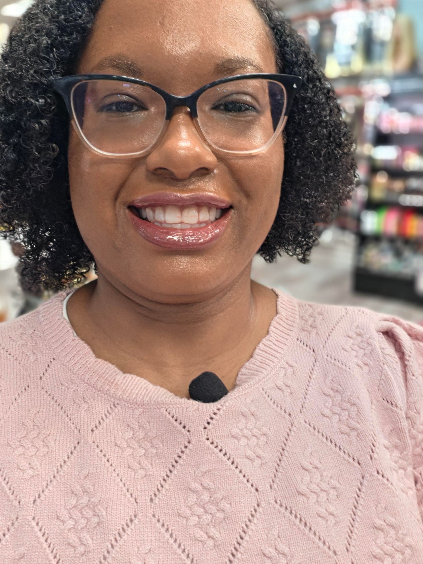 Smiling person wearing glasses and a pink sweater in a store aisle.