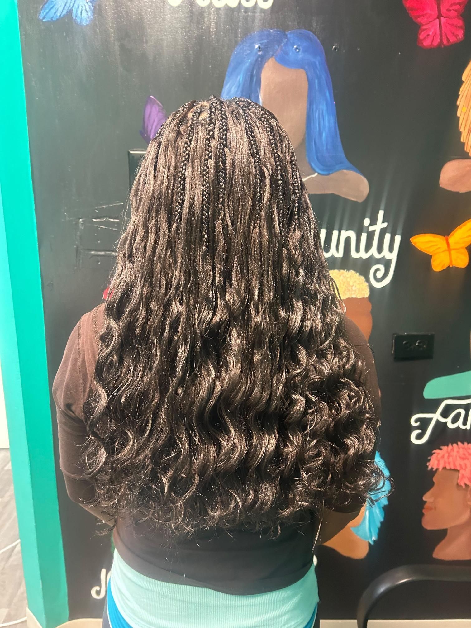 Person with long wavy braids standing before a colorful mural with butterflies and a blue wig display.