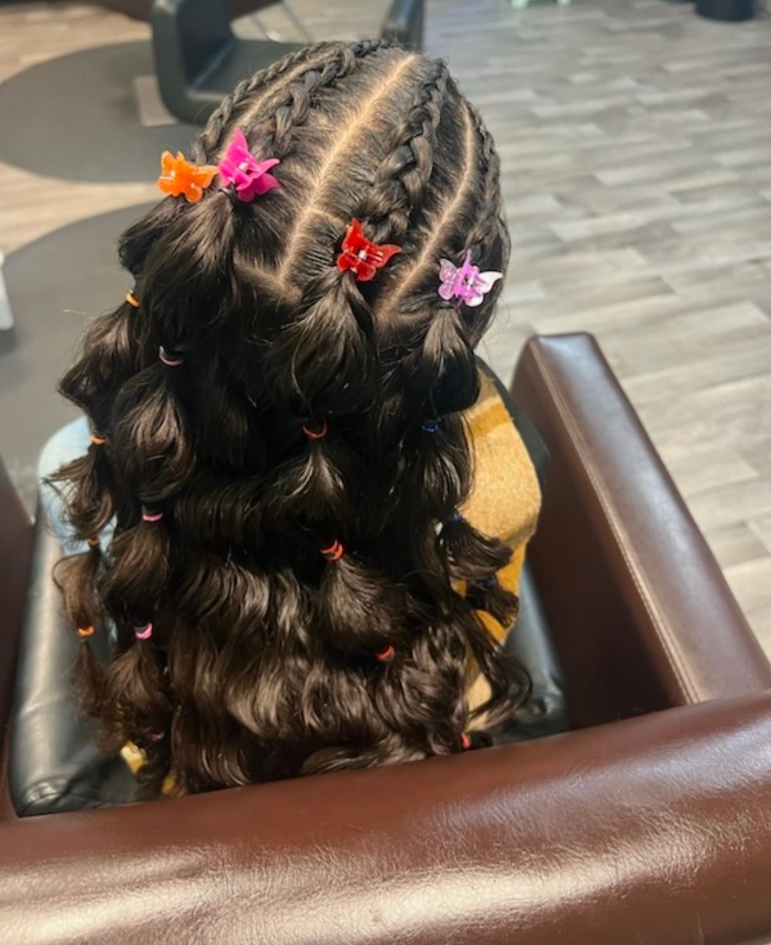 Two braided pigtails with colorful butterfly clips on long wavy dark hair, viewed from behind in a salon chair