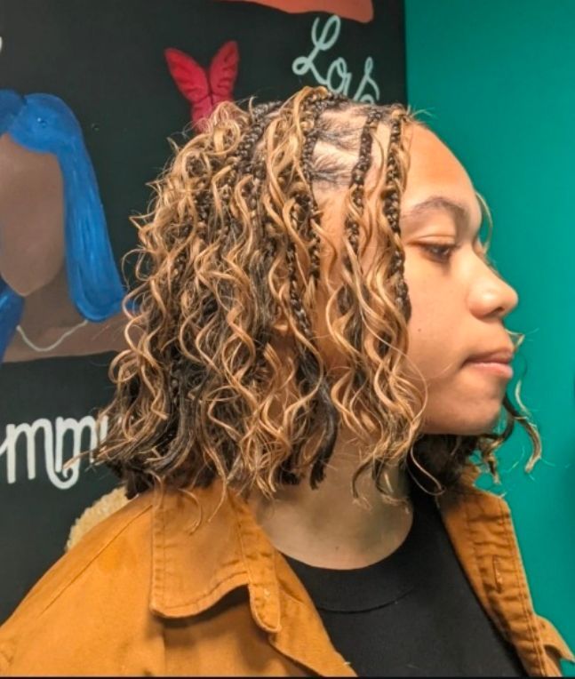 Side profile of a person with shoulder-length curly hair, wearing a mustard jacket in front of a teal wall.