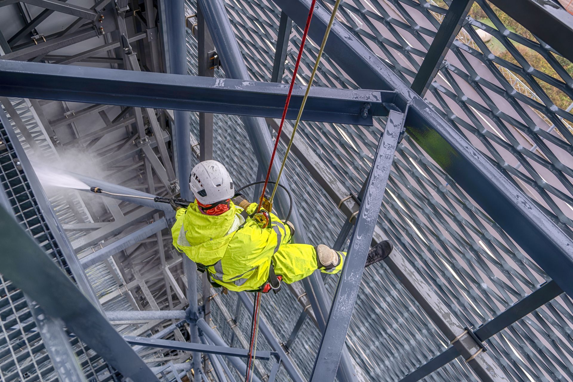 A worker in safety gear rappels, pressure washing a metal structure.