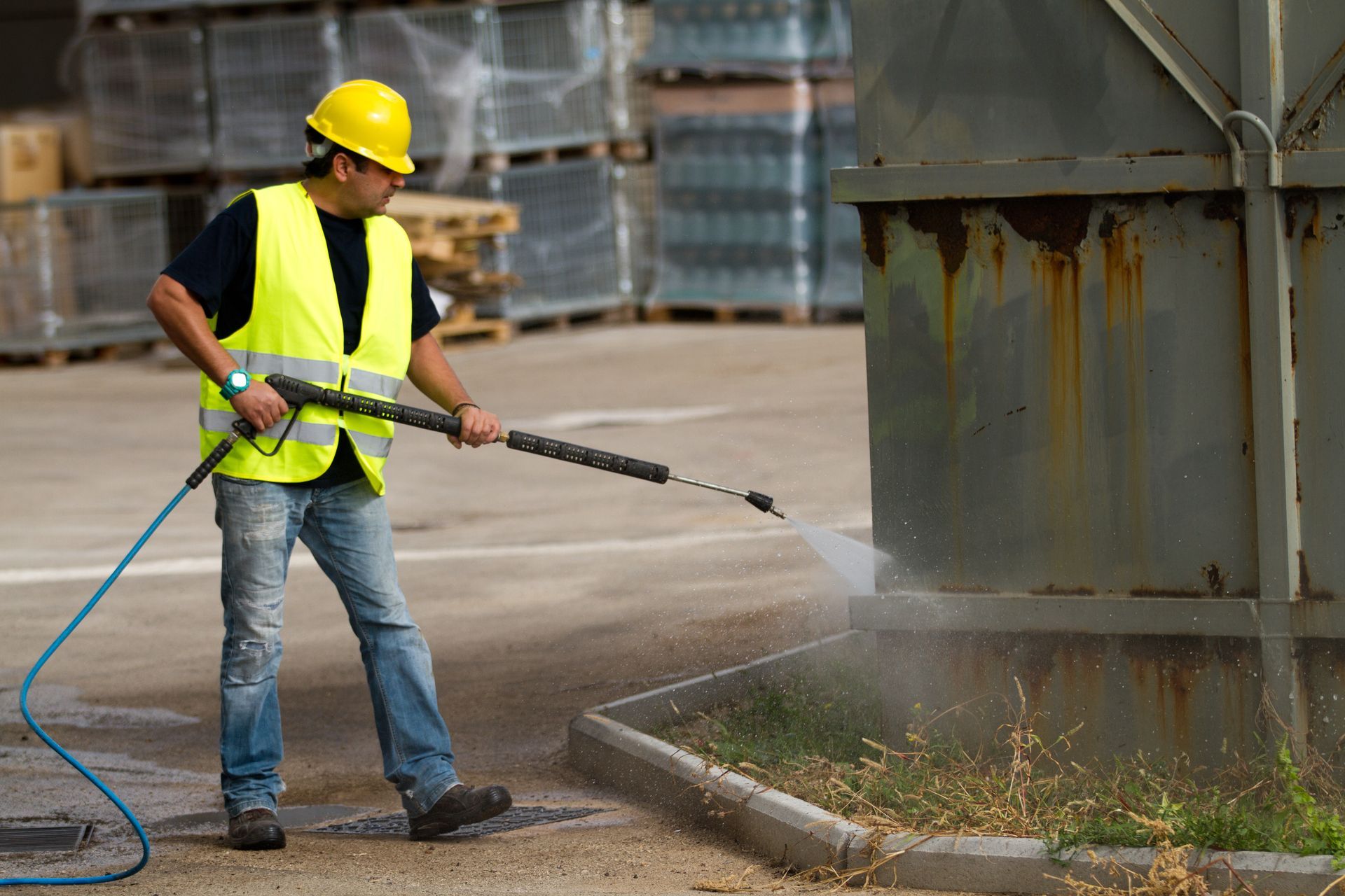 Worker in safety vest power washing a rusty metal container, outdoors.