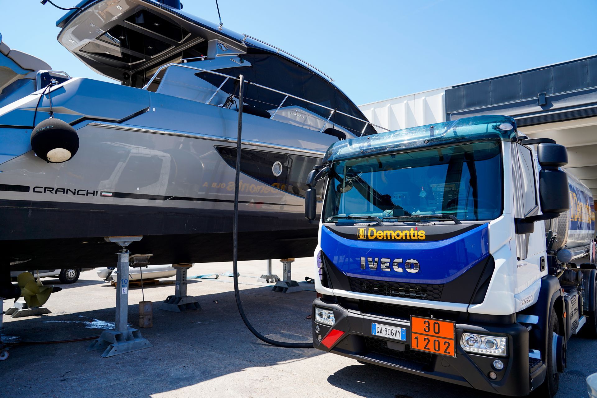 Un camion blu e bianco è affiancato a un grande yacht su un rimorchio in un porto turistico.