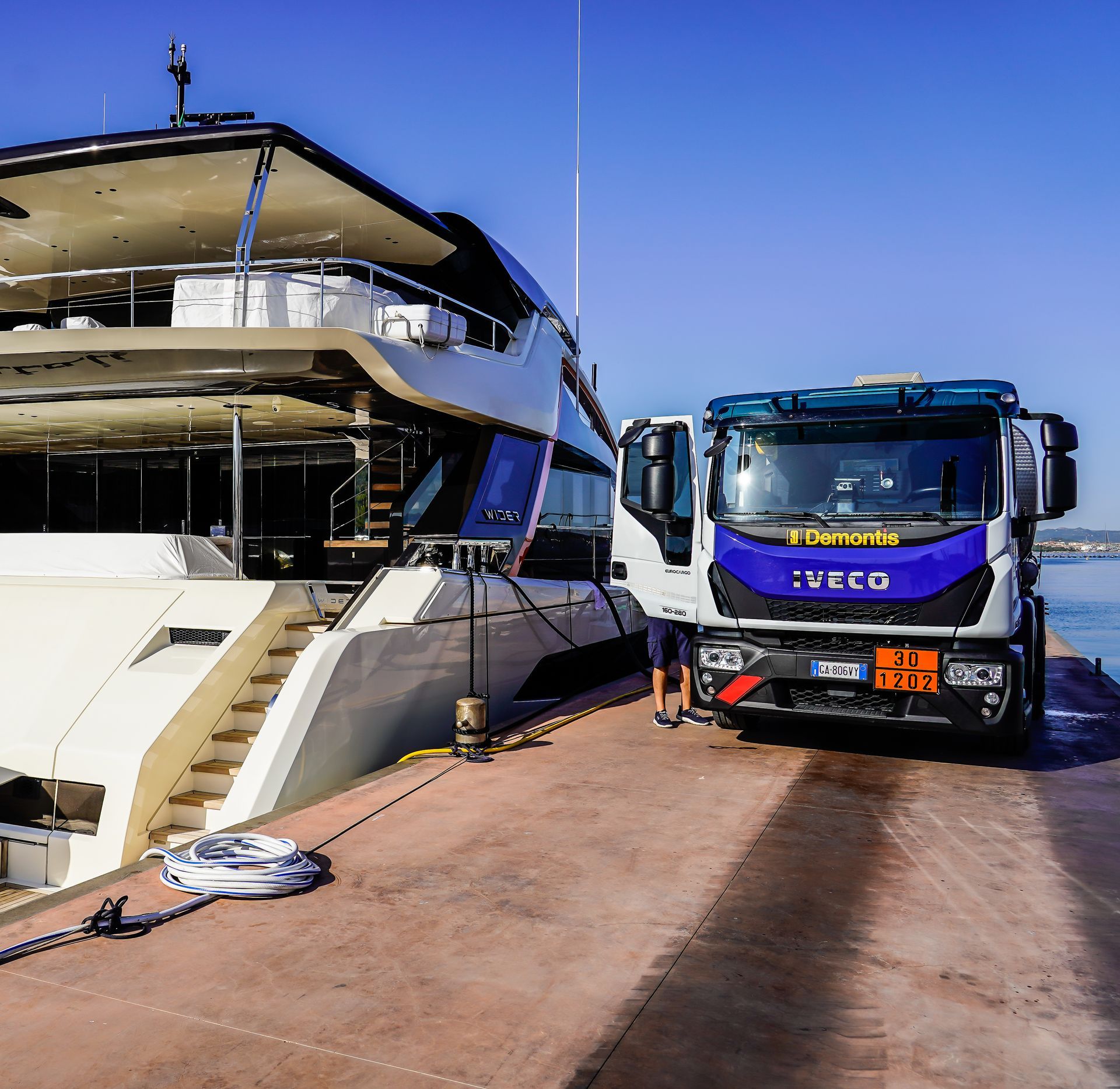 Uno yacht bianco ormeggiato accanto a un camion IVECO blu su un molo soleggiato.