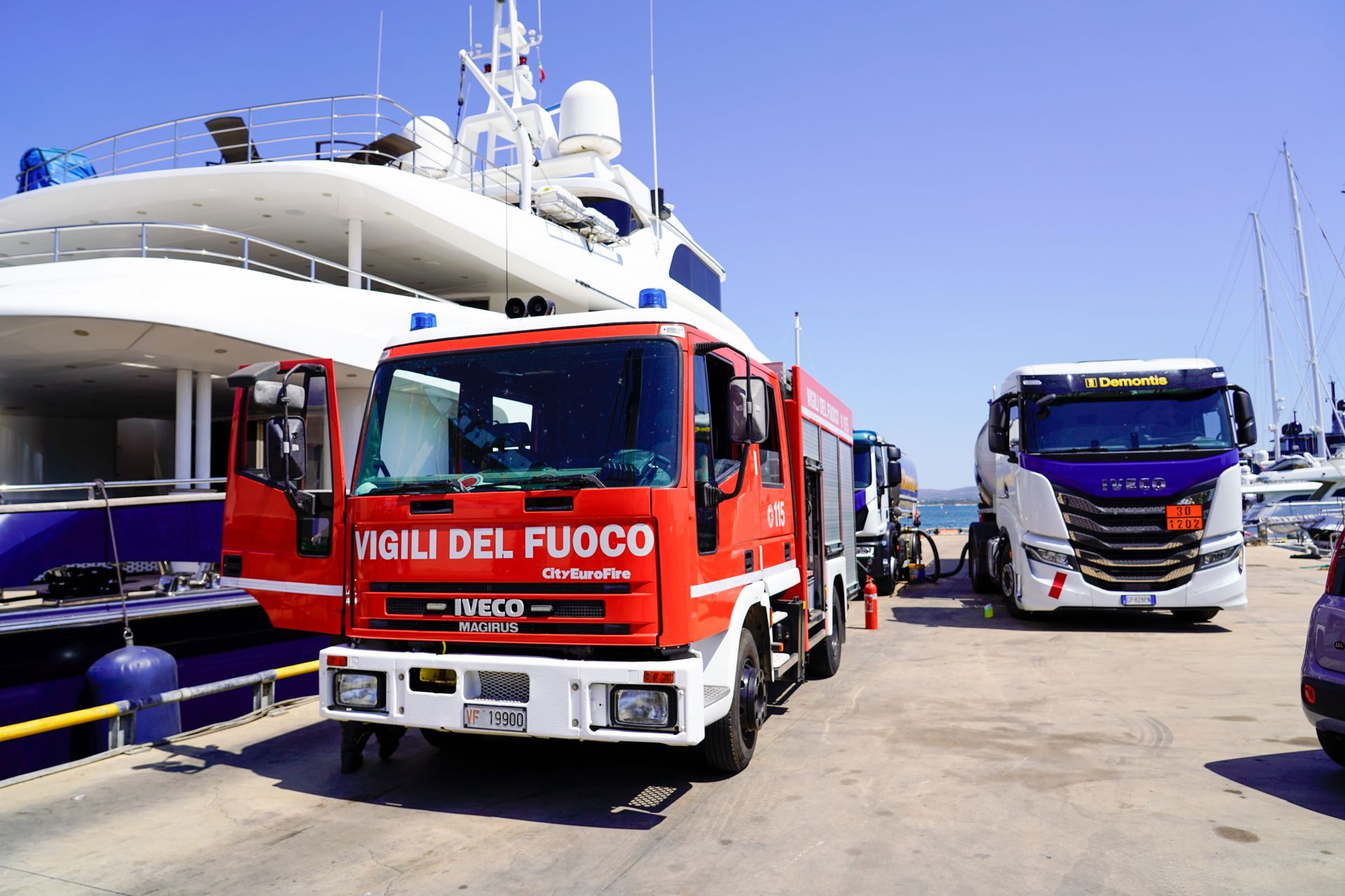 Un camion dei pompieri rosso in porto, accanto a uno yacht, con un autobus blu sullo sfondo.