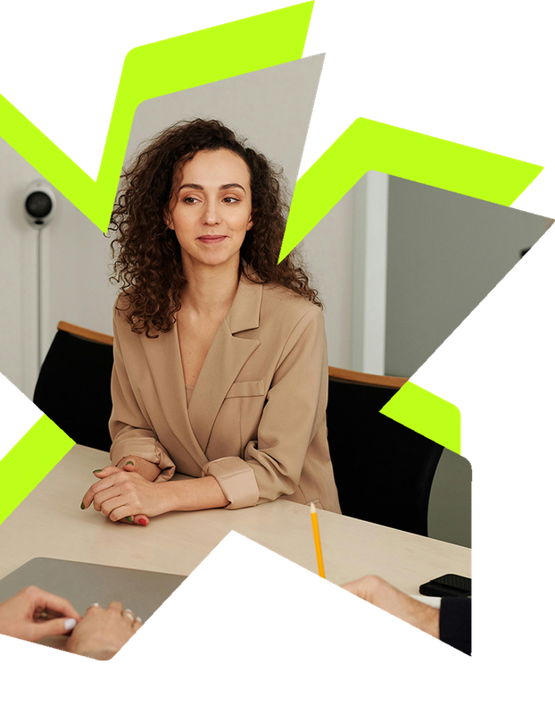 A professional woman in a tan blazer sits at a desk during a meeting, with hands visible in the foreground.