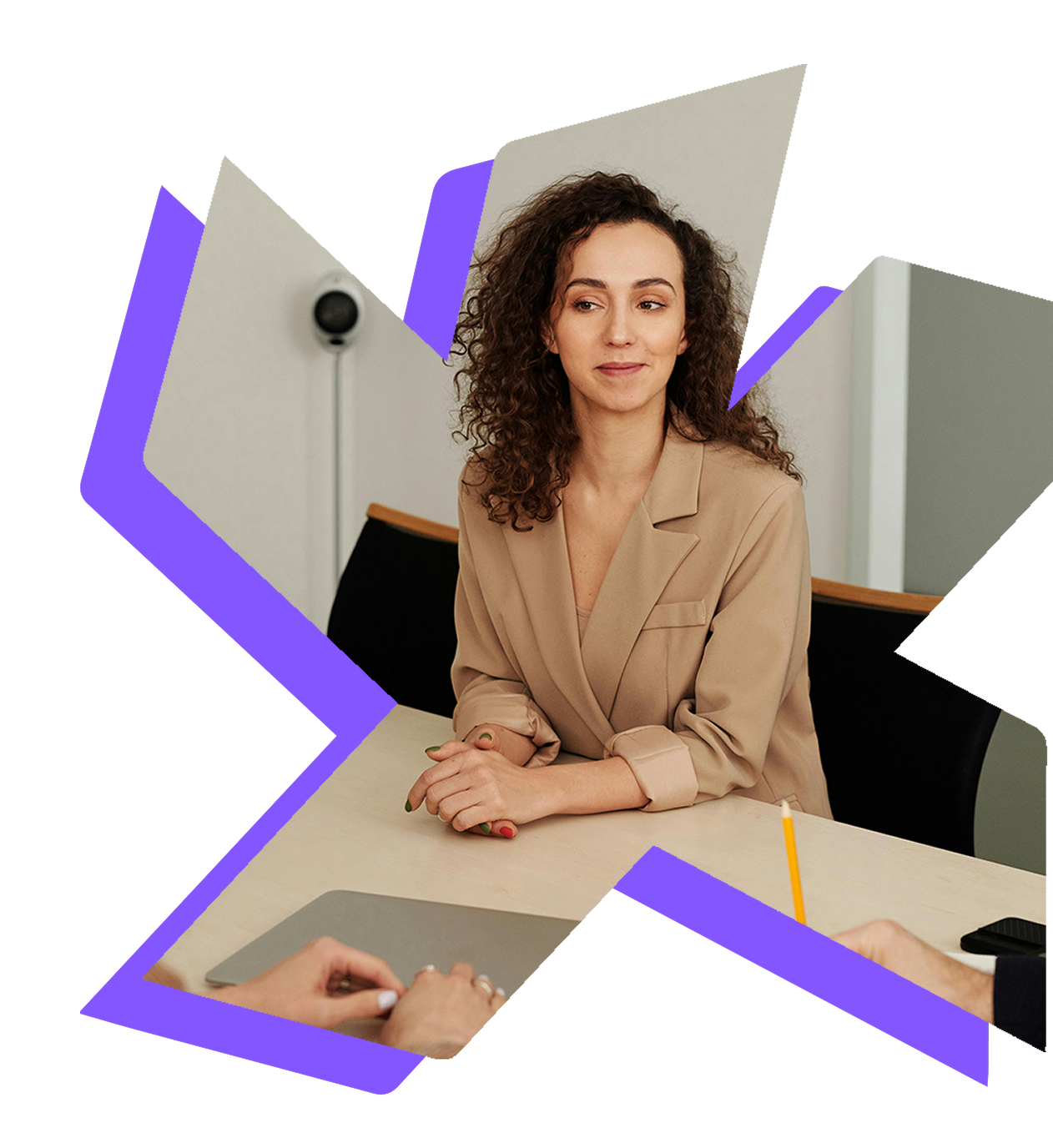 A person with curly hair in a tan blazer sits at a table, participating in a meeting with others partially visible.
