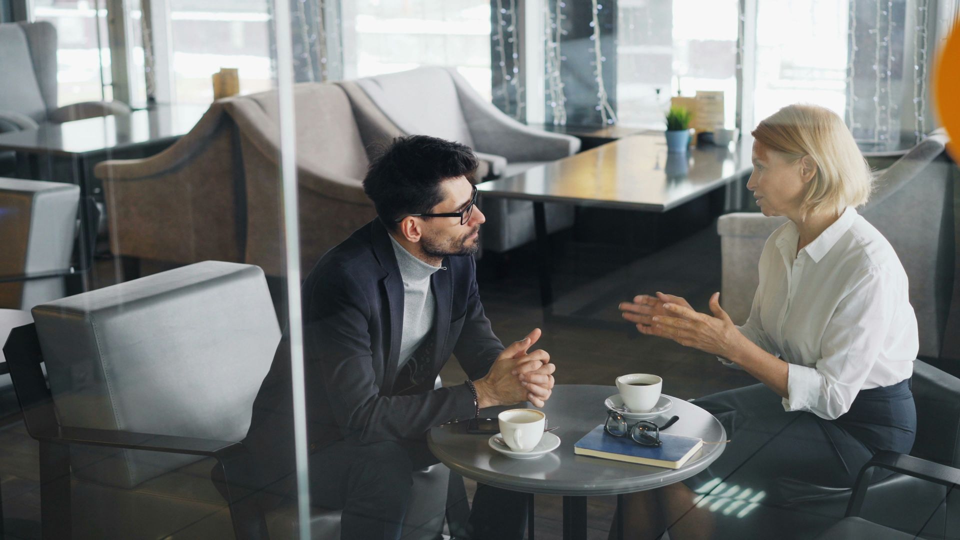 Two people in business attire sit at a small table, talking over coffee in a cafe.