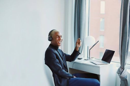 Person with headphones, smiling, waving at laptop near window.