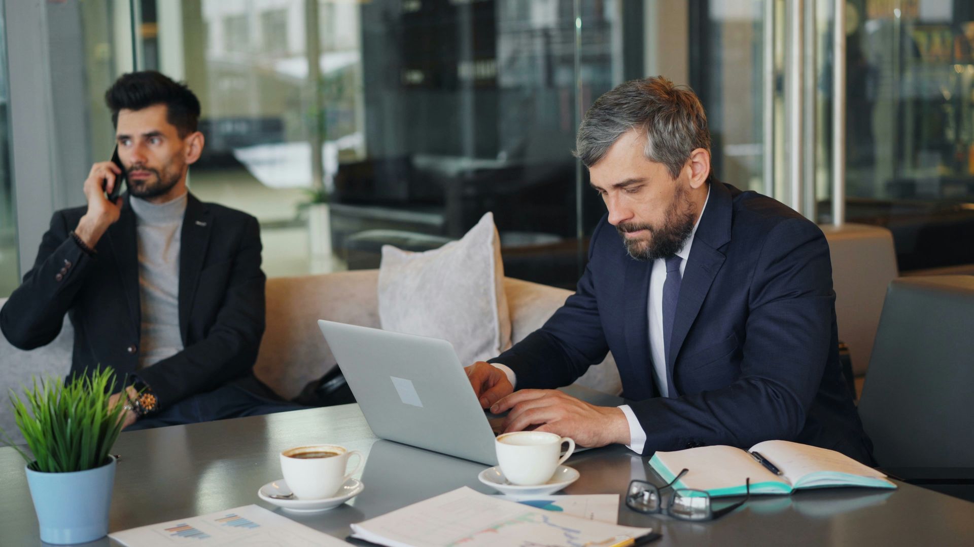 Two men in suits, one on phone, the other working on a laptop at a table with coffee cups and documents.