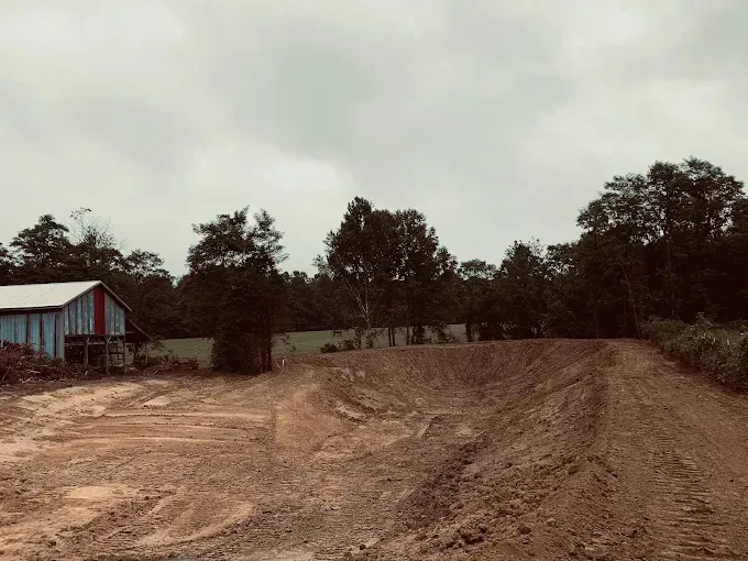 Earthen motocross track, barn, trees under overcast sky. Brown soil, blue and red barn.