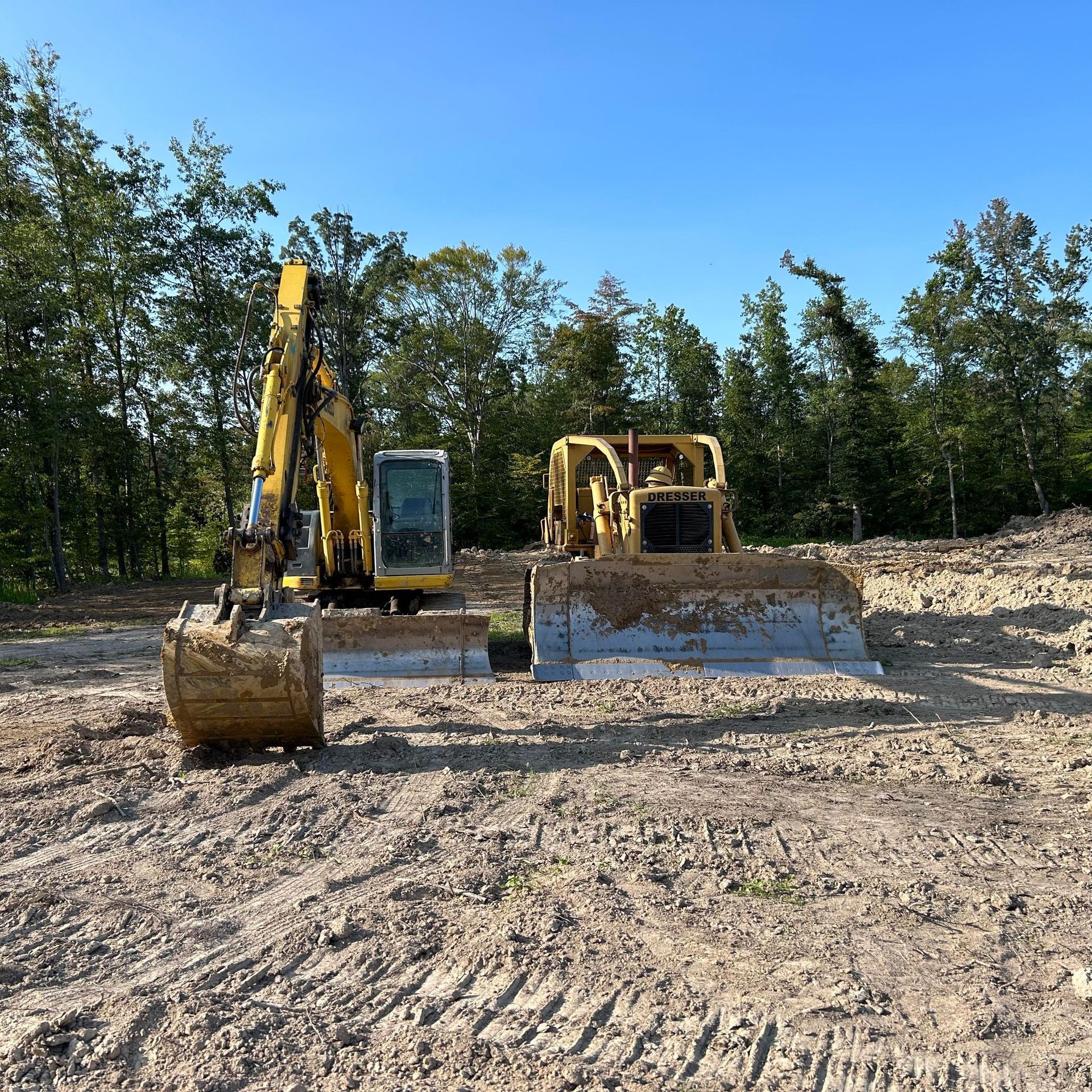 Excavator and bulldozer on dirt. Blue sky, trees in background. Heavy machinery work site.