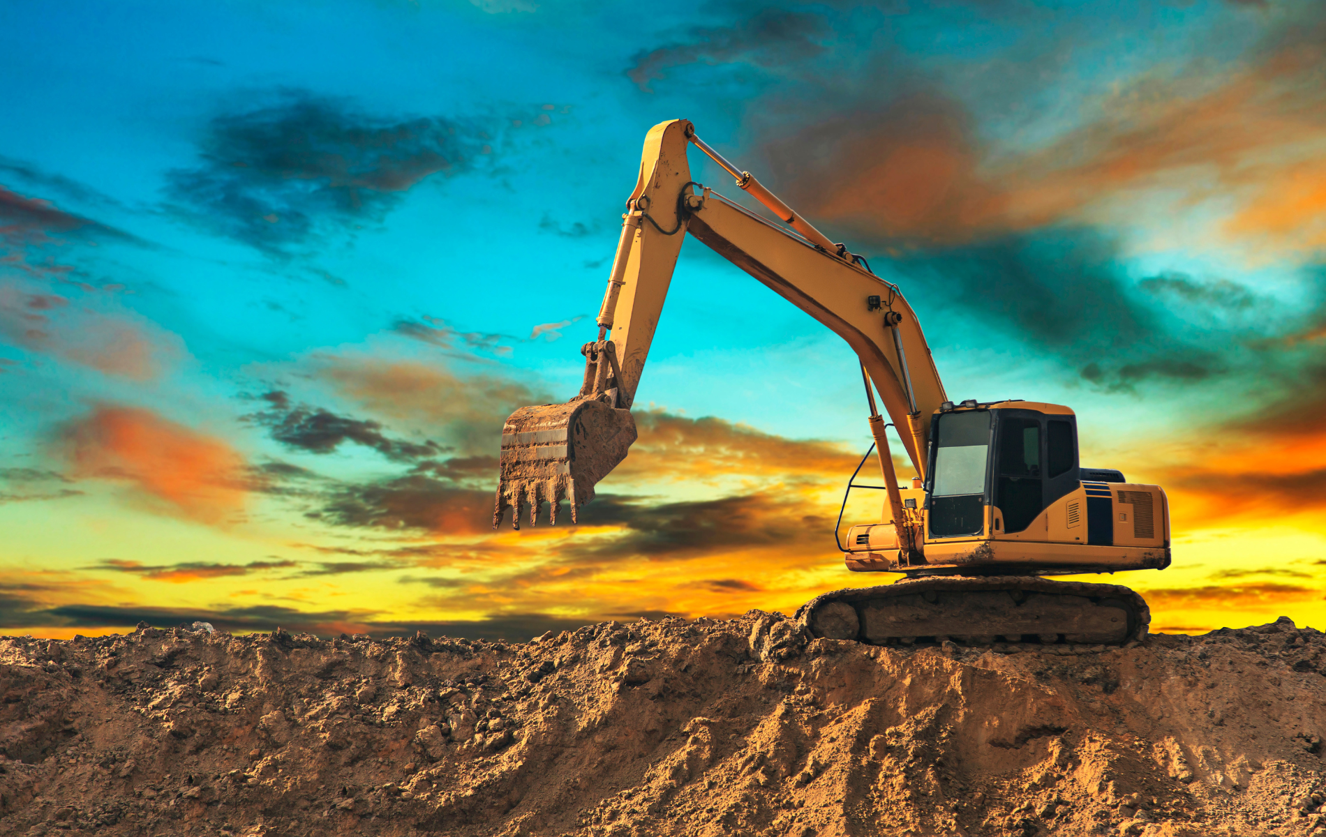 Yellow excavator on a dirt mound against a colorful sunset sky.