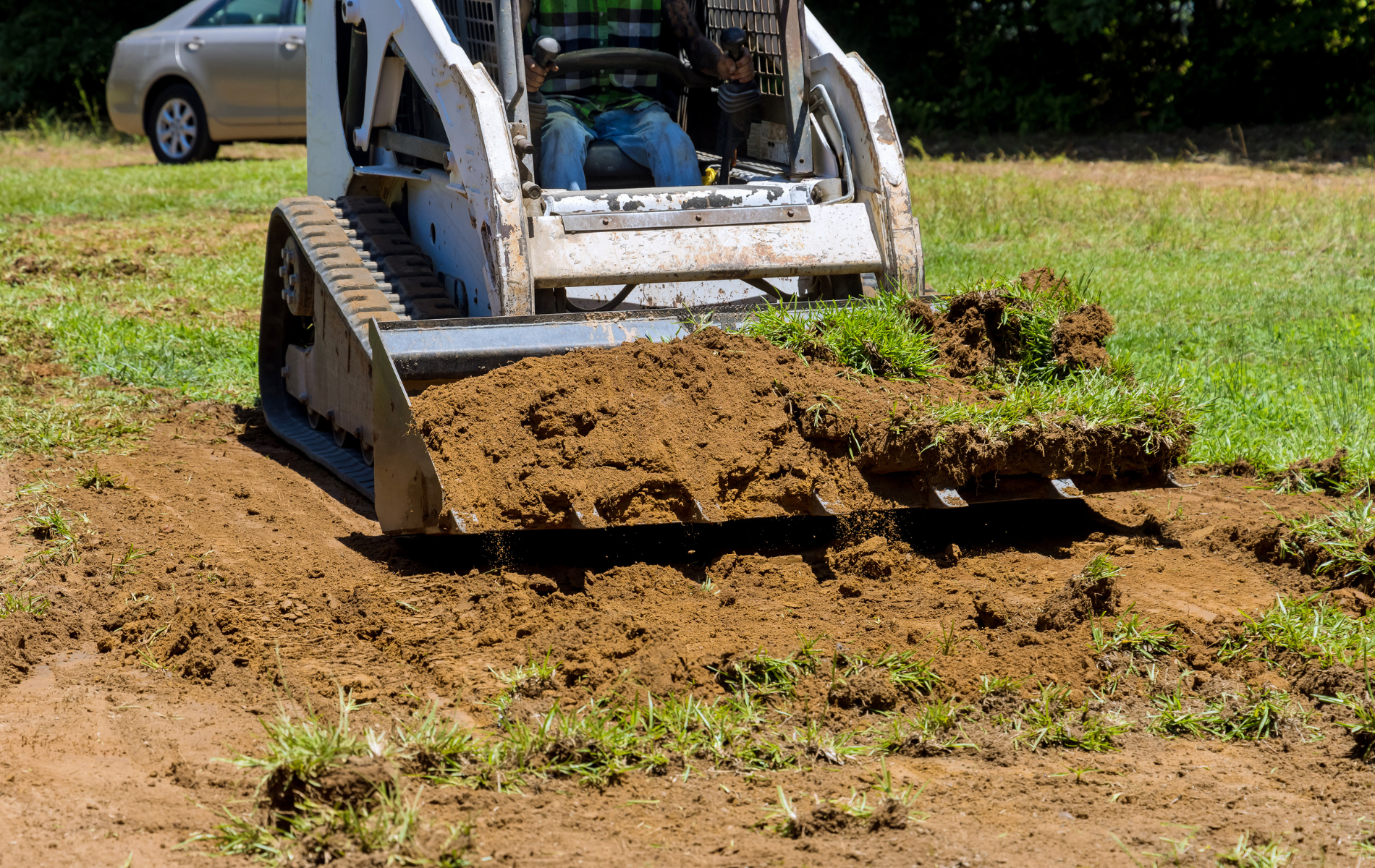 Bobcat removing dirt and grass from a grassy area. A car is visible in the background.