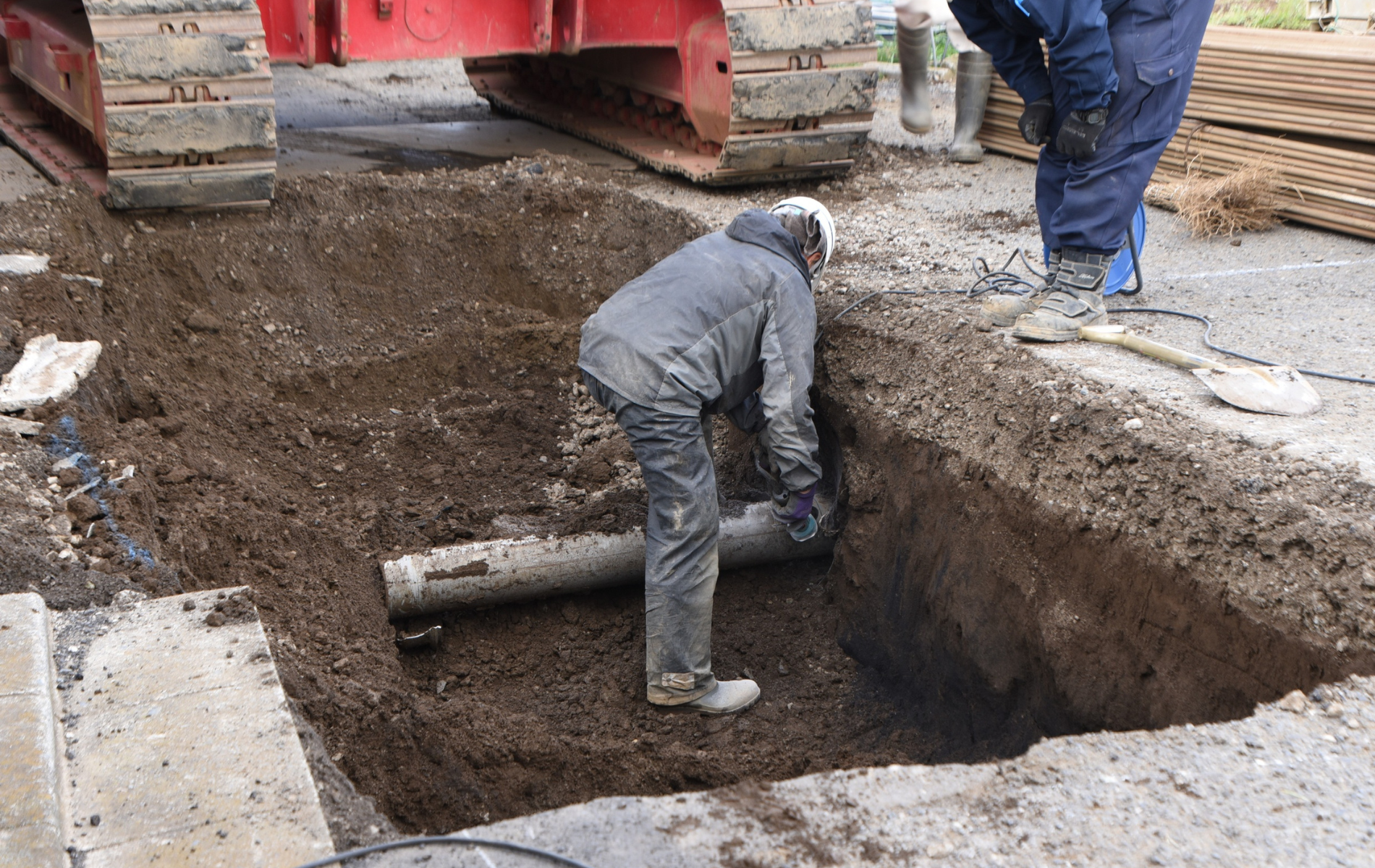 Construction workers installing a pipe in a trench next to a road, with heavy machinery visible.