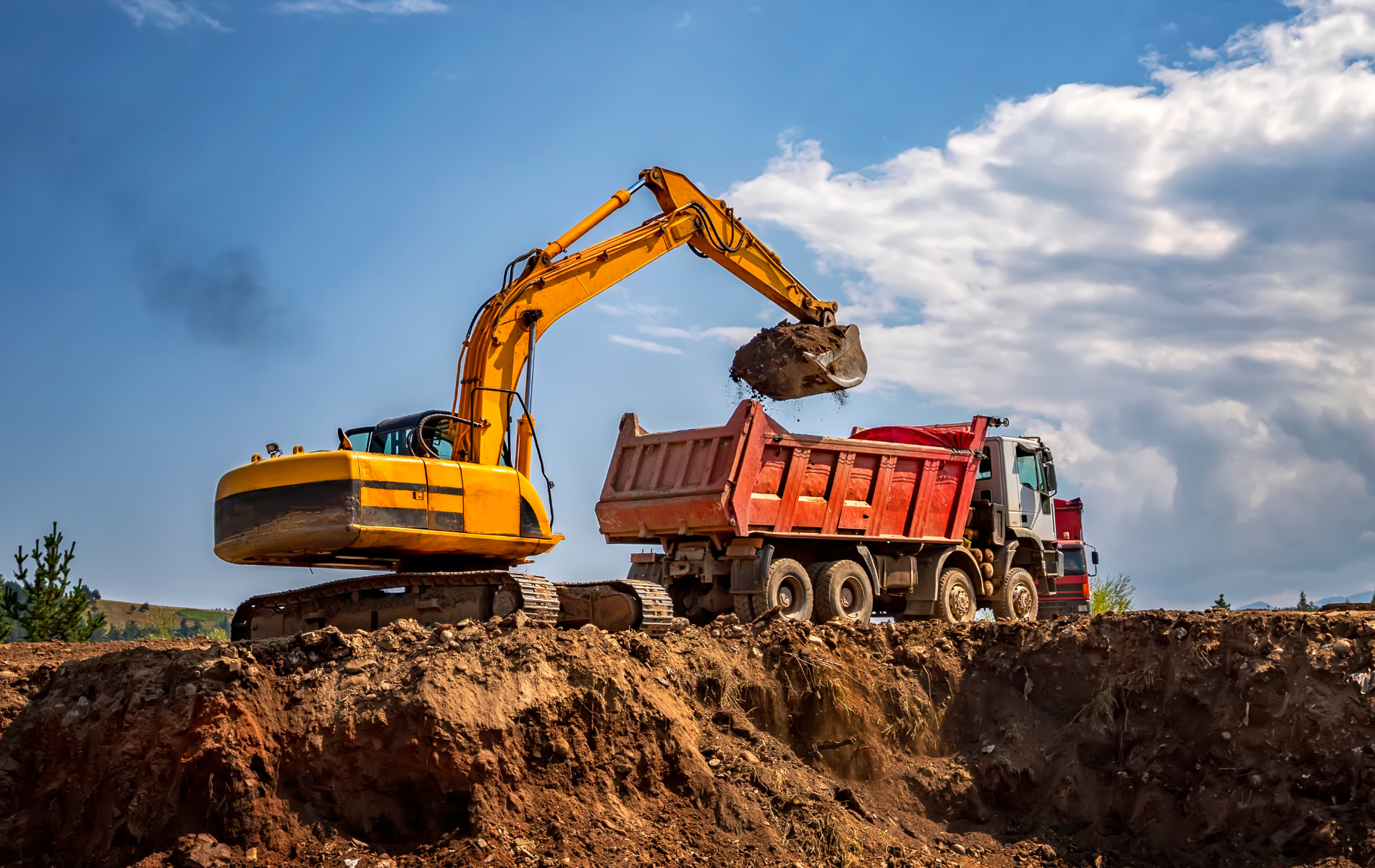 Yellow excavator loading dirt into a red dump truck, under a blue sky.