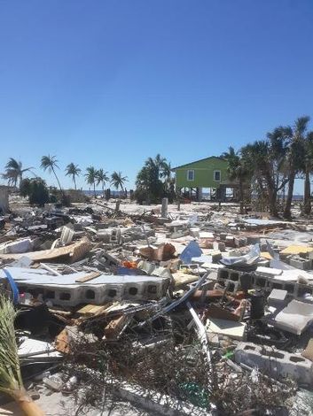 Debris field after a hurricane on a beach, with a green house standing in the distance.