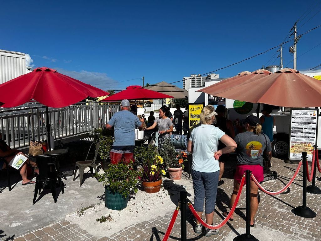 People line up at a food truck under red and tan umbrellas on a sunny day.