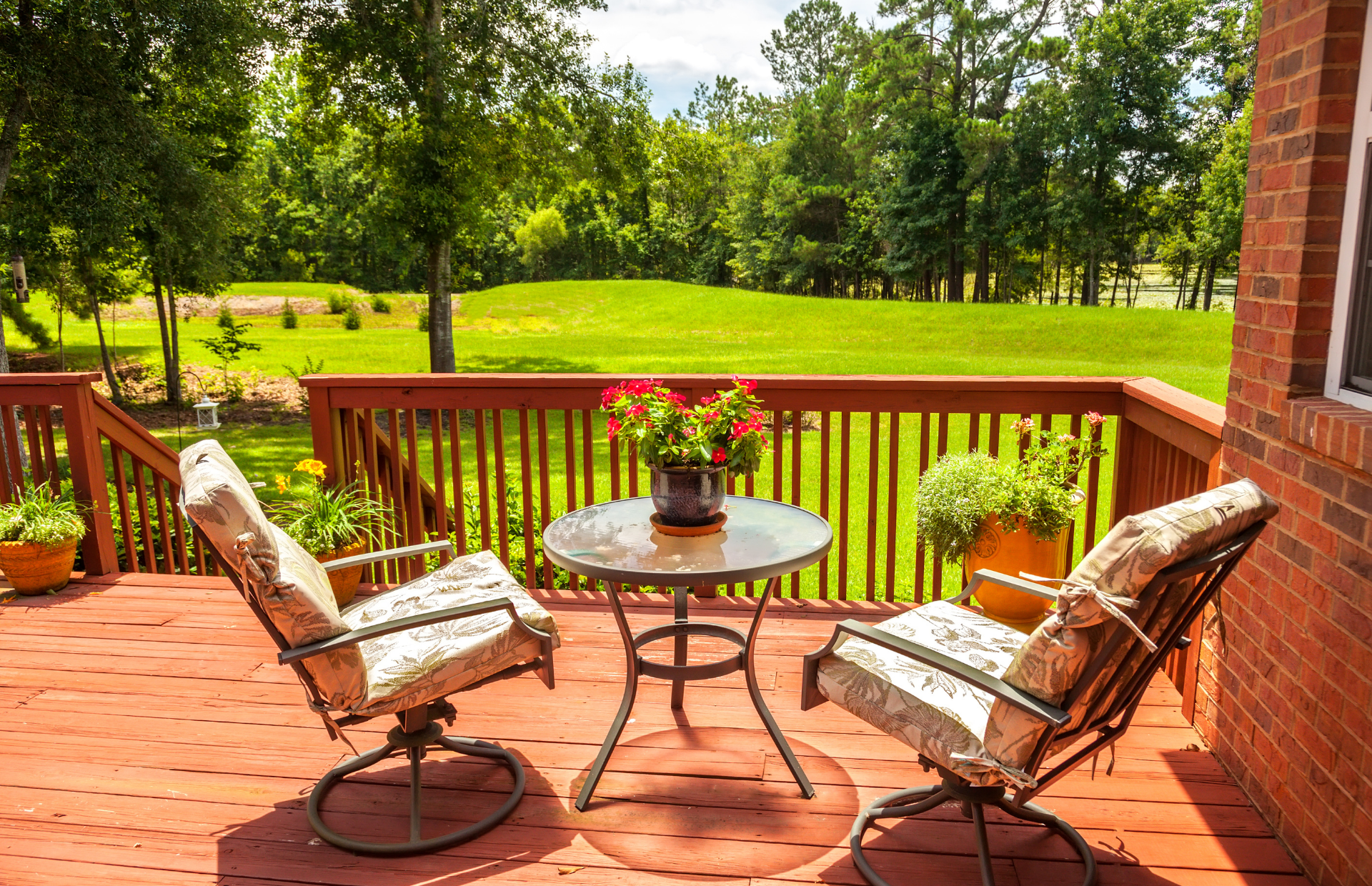 Patio with two cushioned chairs, round table, and flower pot overlooking a green backyard and trees