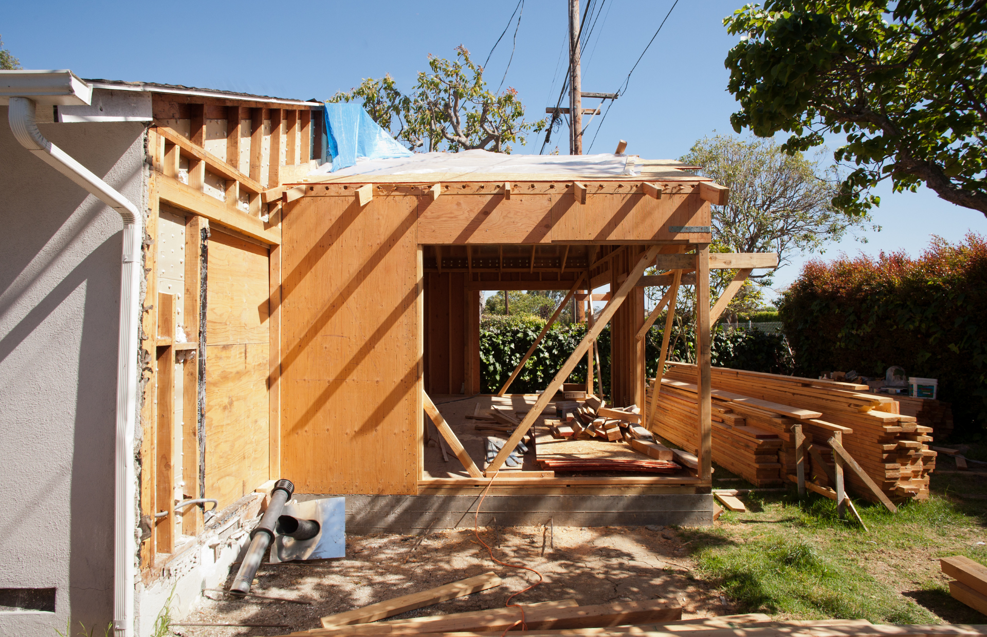 Wood-framed house addition under construction with open beams and tools in a sunny yard