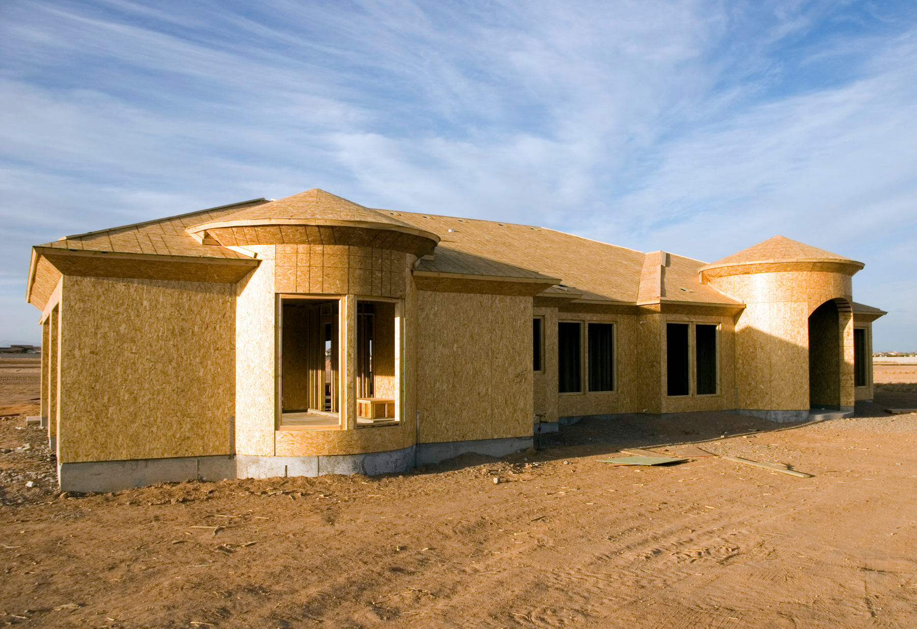 Single-story tan house under construction on a barren lot beneath a blue sky