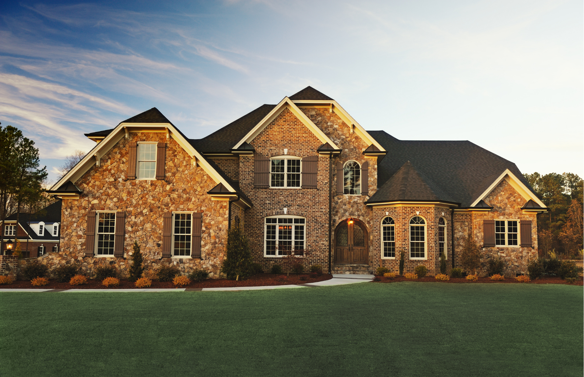 Large stone suburban house with a dark roof and manicured green lawn at sunset