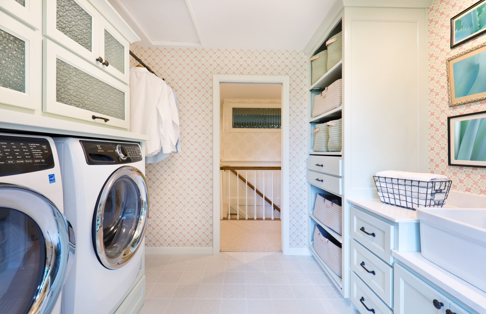 Bright laundry room with front-loading washer, dryer, white cabinetry, and patterned wallpaper