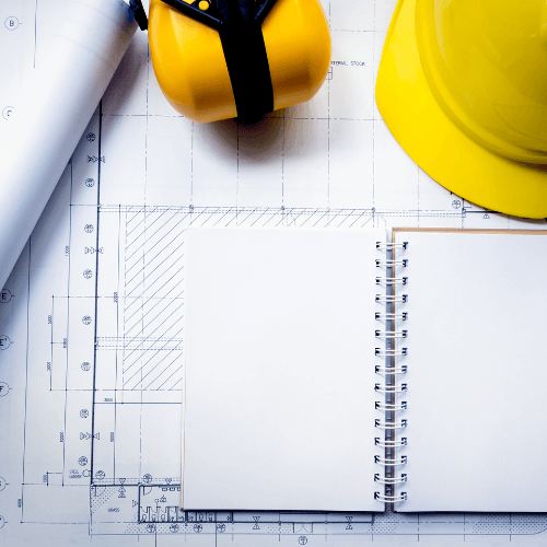 Construction plans with yellow hard hat, earmuffs, and open spiral notebook on blueprints