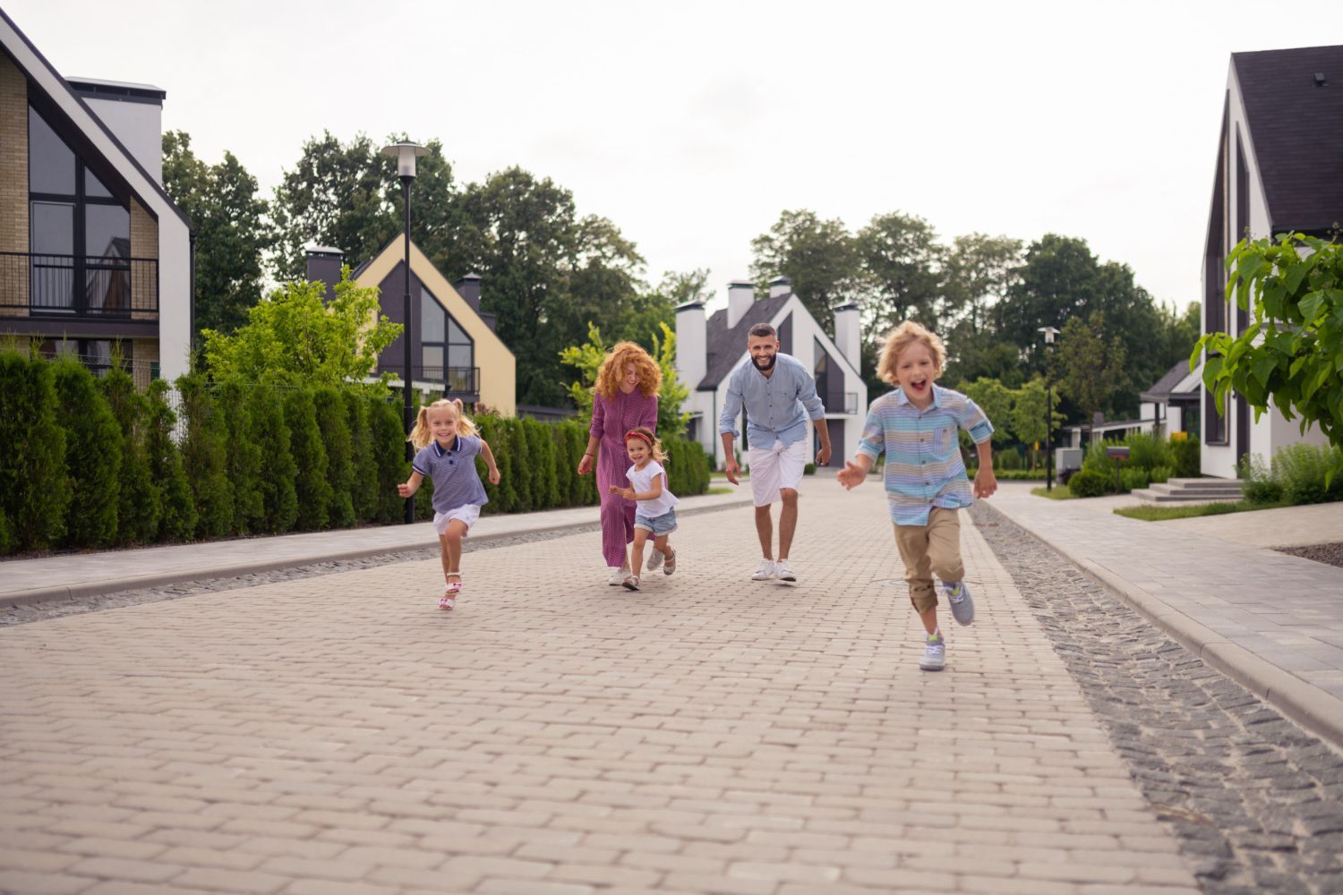 Family of five running down a brick-paved street past modern homes; two children lead the way.