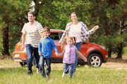 Family running happily towards the camera in a grassy field, red car in the background.