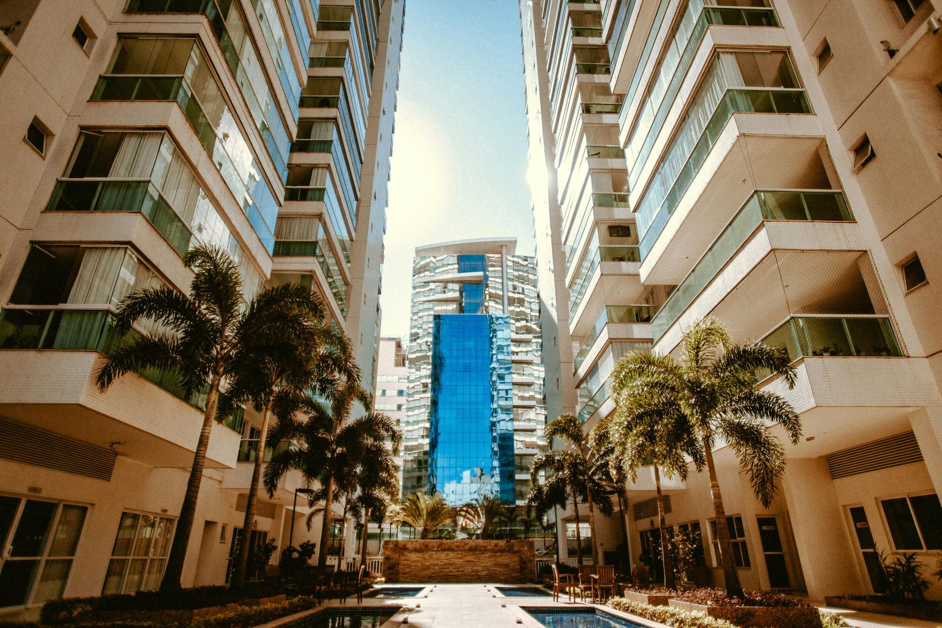 Tall beige apartment buildings frame a glass skyscraper, with palm trees and a small pool in the foreground.