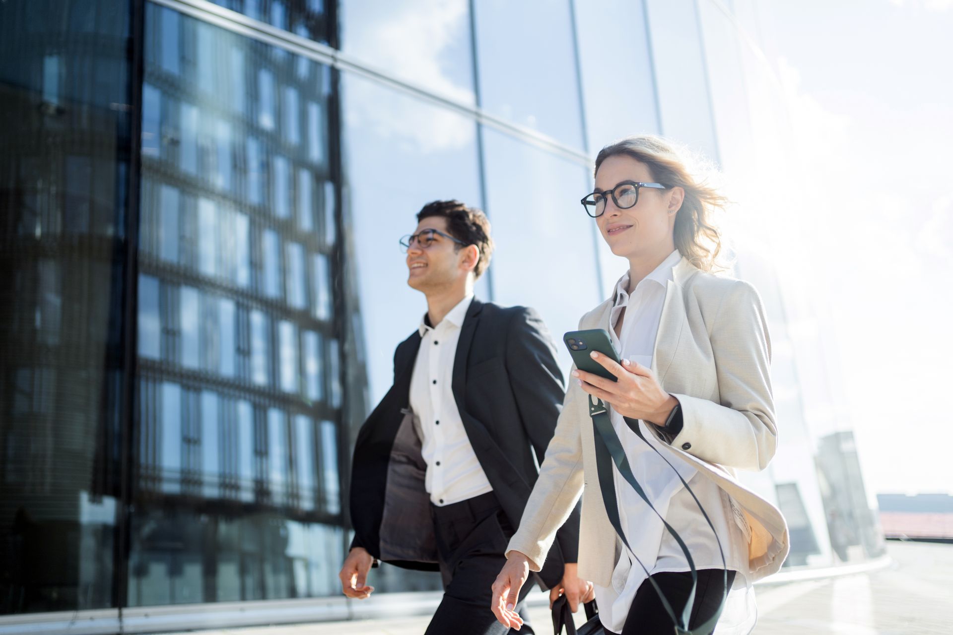 Two businesspeople walk near a modern building; the woman holds a phone, the man smiles.