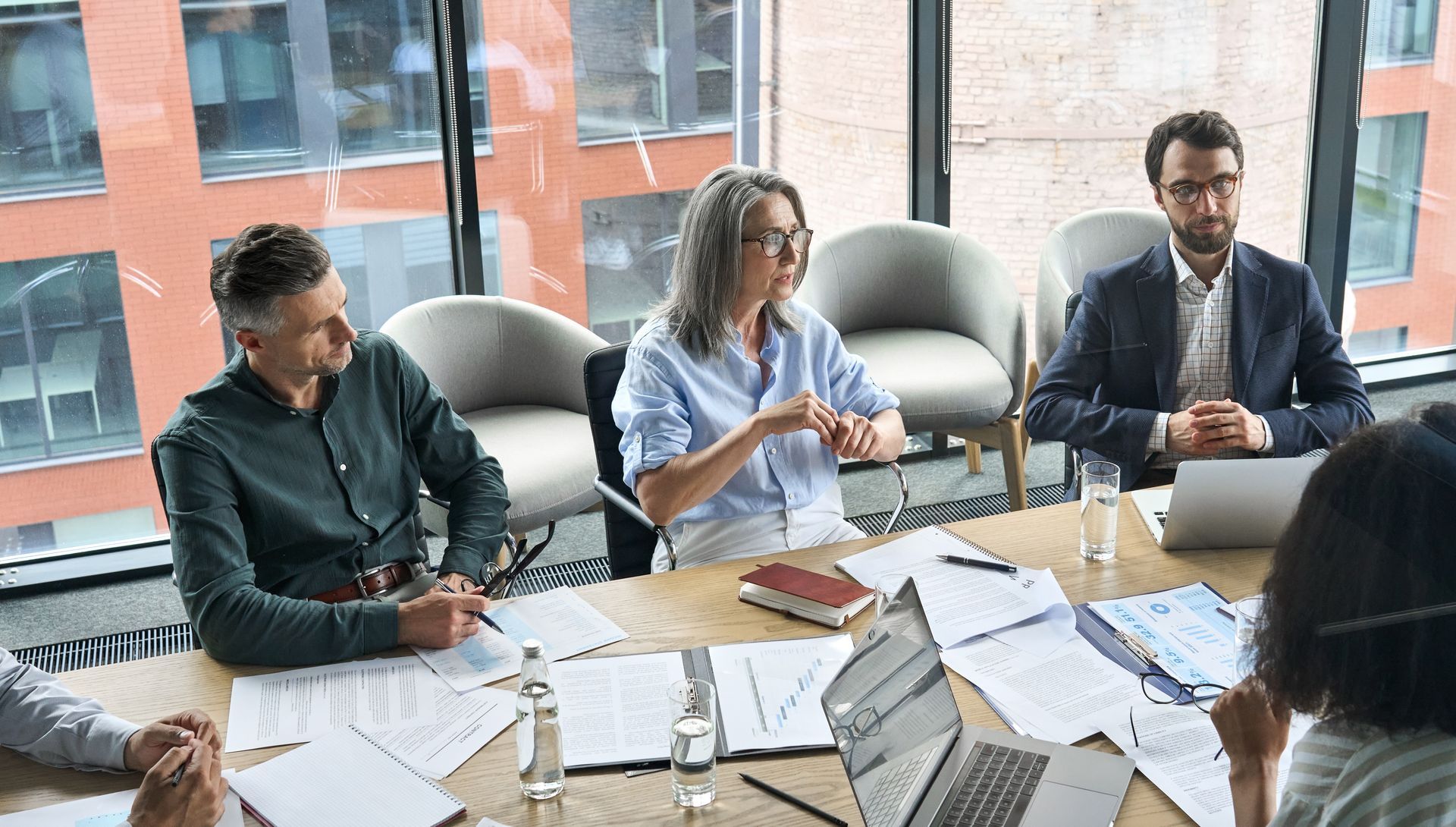 People in a meeting at a table with papers and laptops, discussing ideas in an office with a window.