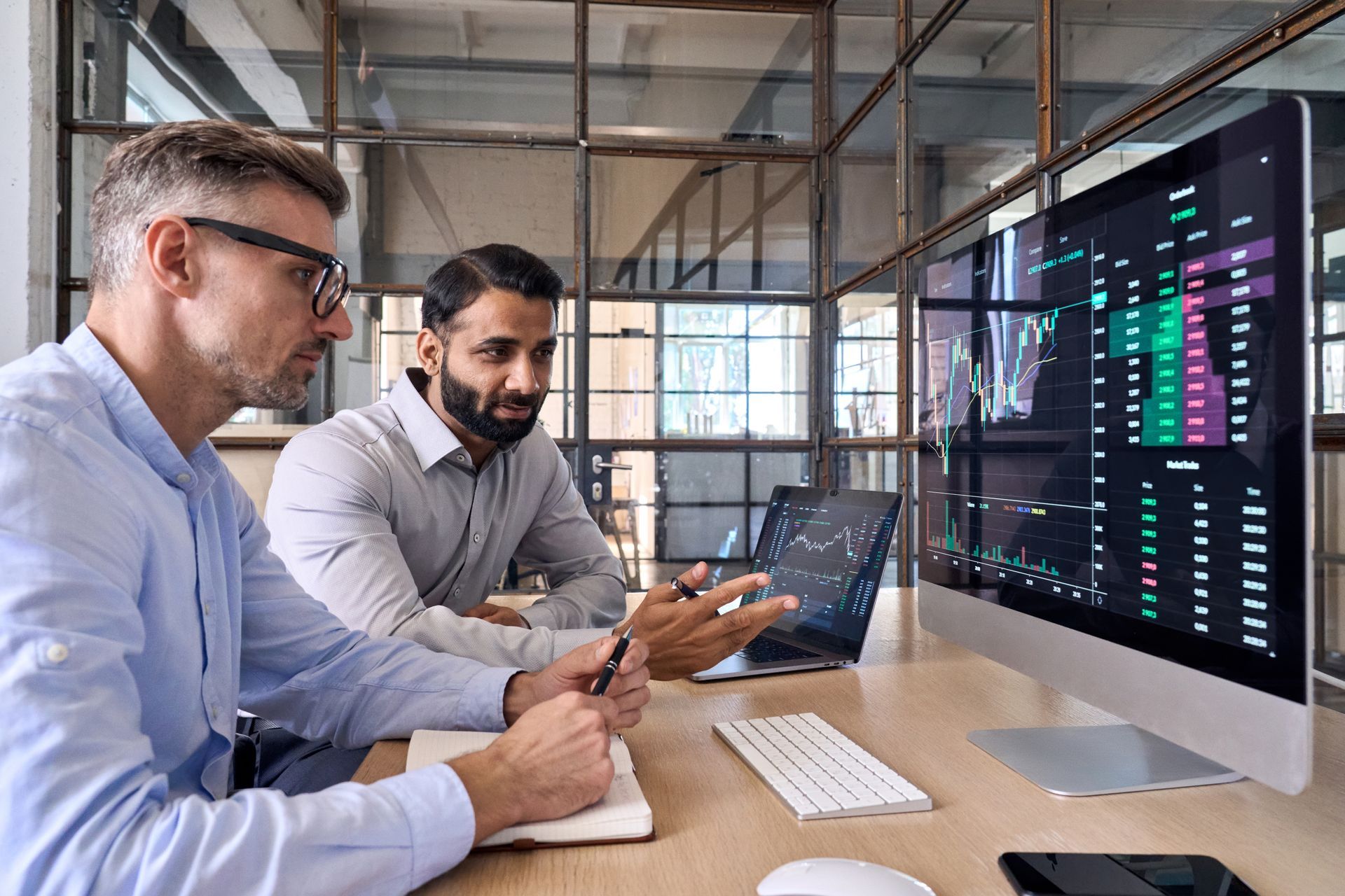 Two men in an office looking at stock charts on a monitor and laptop, discussing investments.