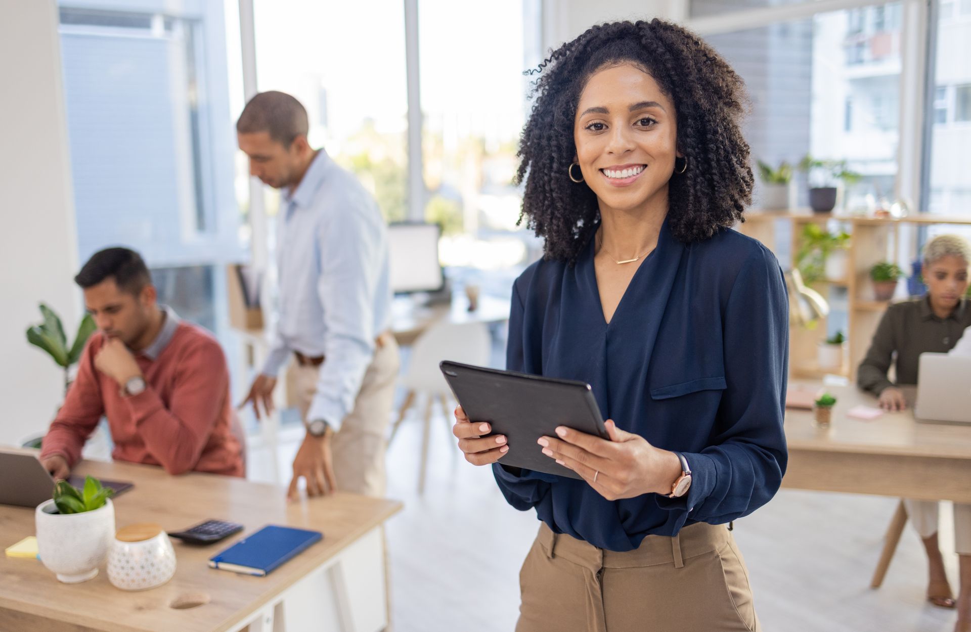Woman holding tablet smiles in a bright office, colleagues work in background.