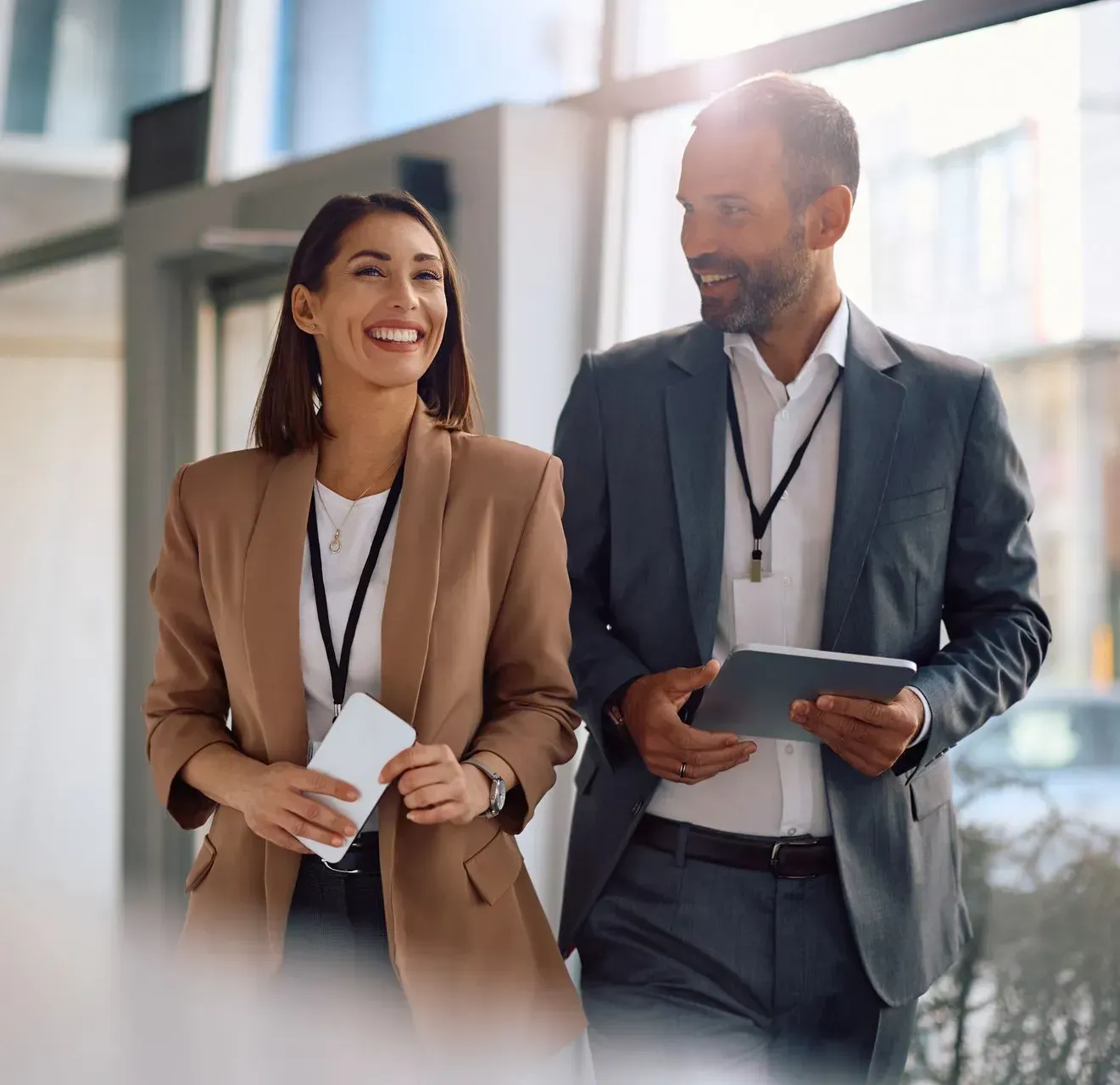 Woman and man in business attire walking and smiling, indoors, holding tablet and phone.