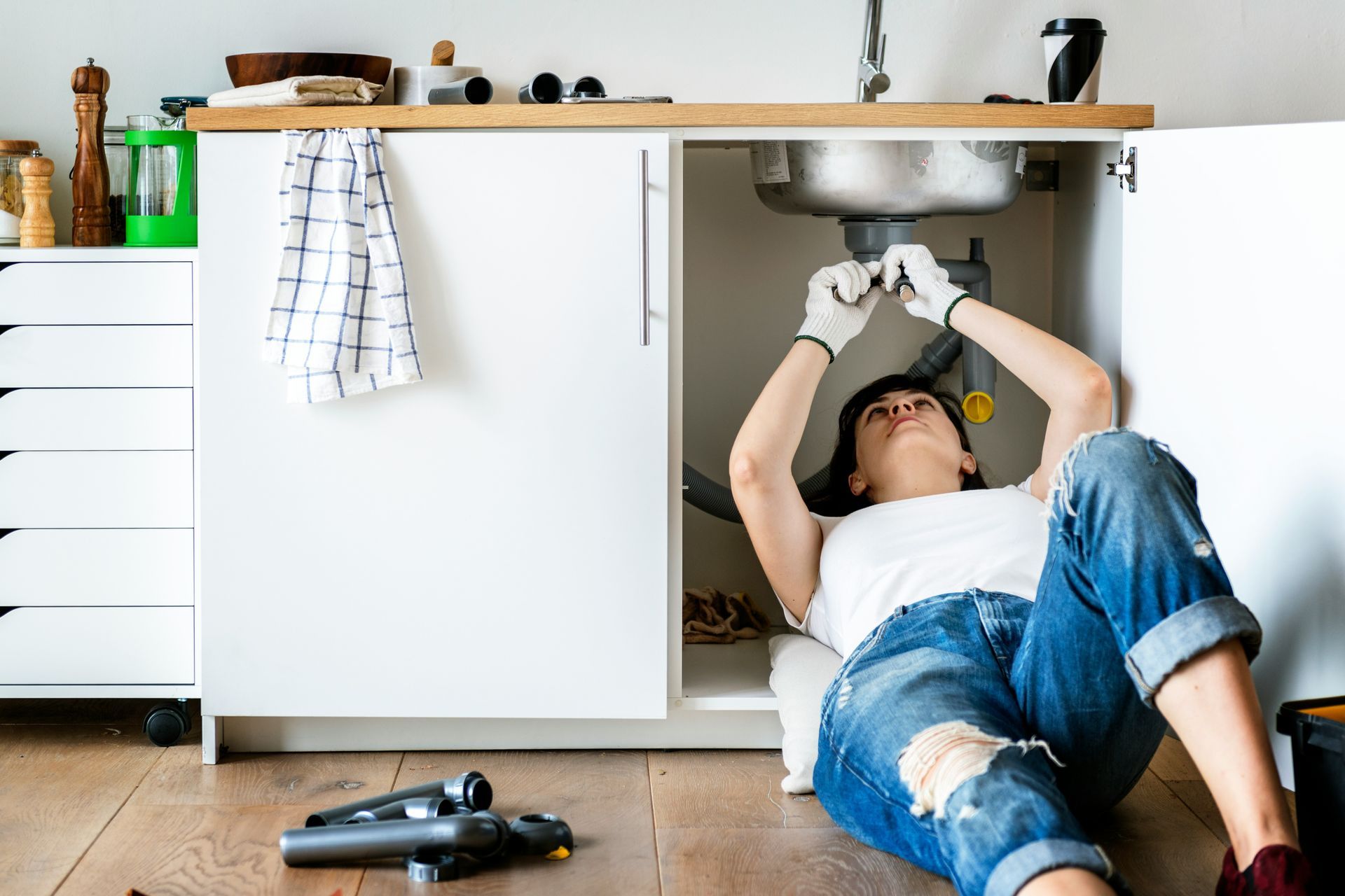 Person lying on kitchen floor under sink, reaching up with tools during repairs.