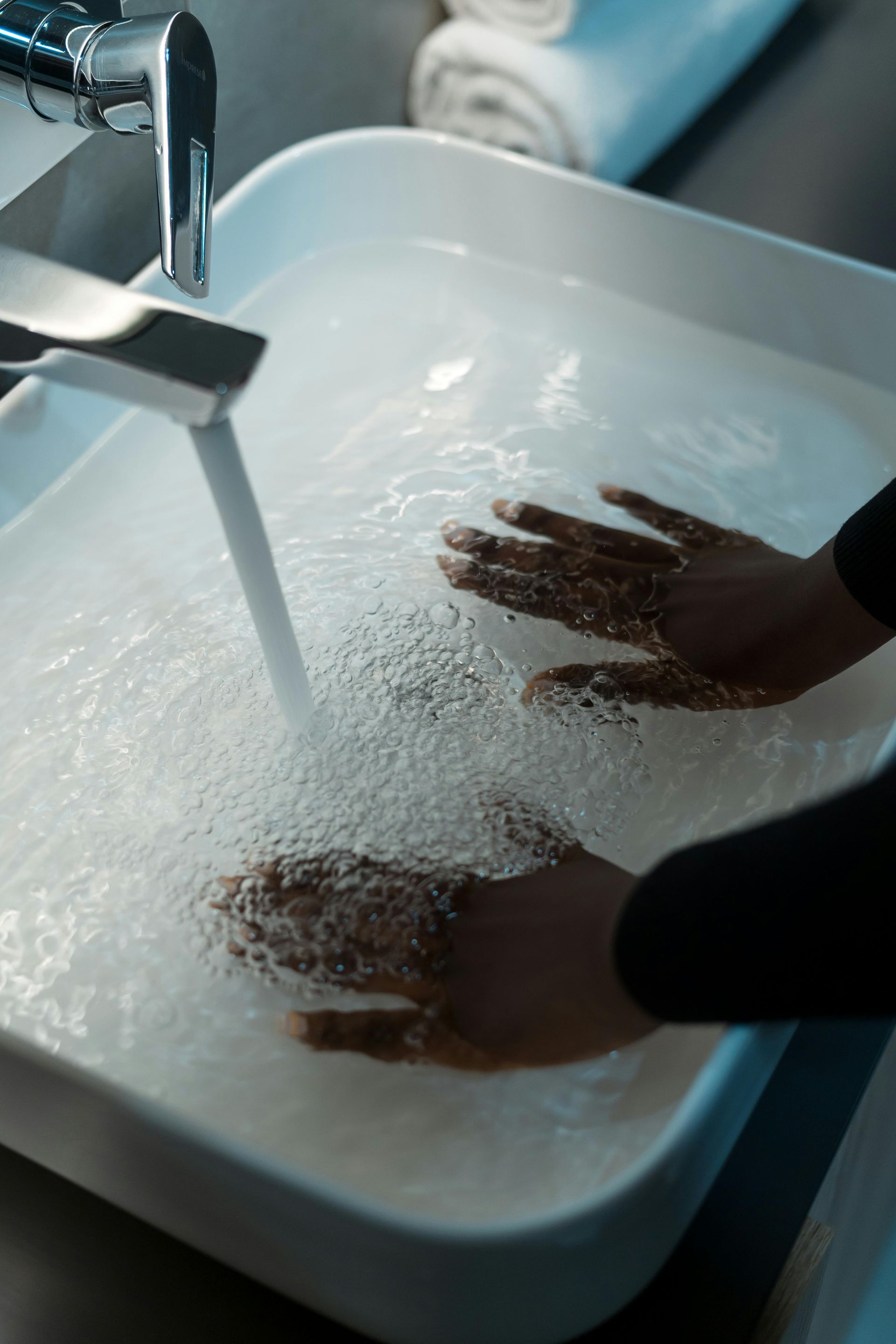 Hands washing under running faucet in a white sink filled with water.