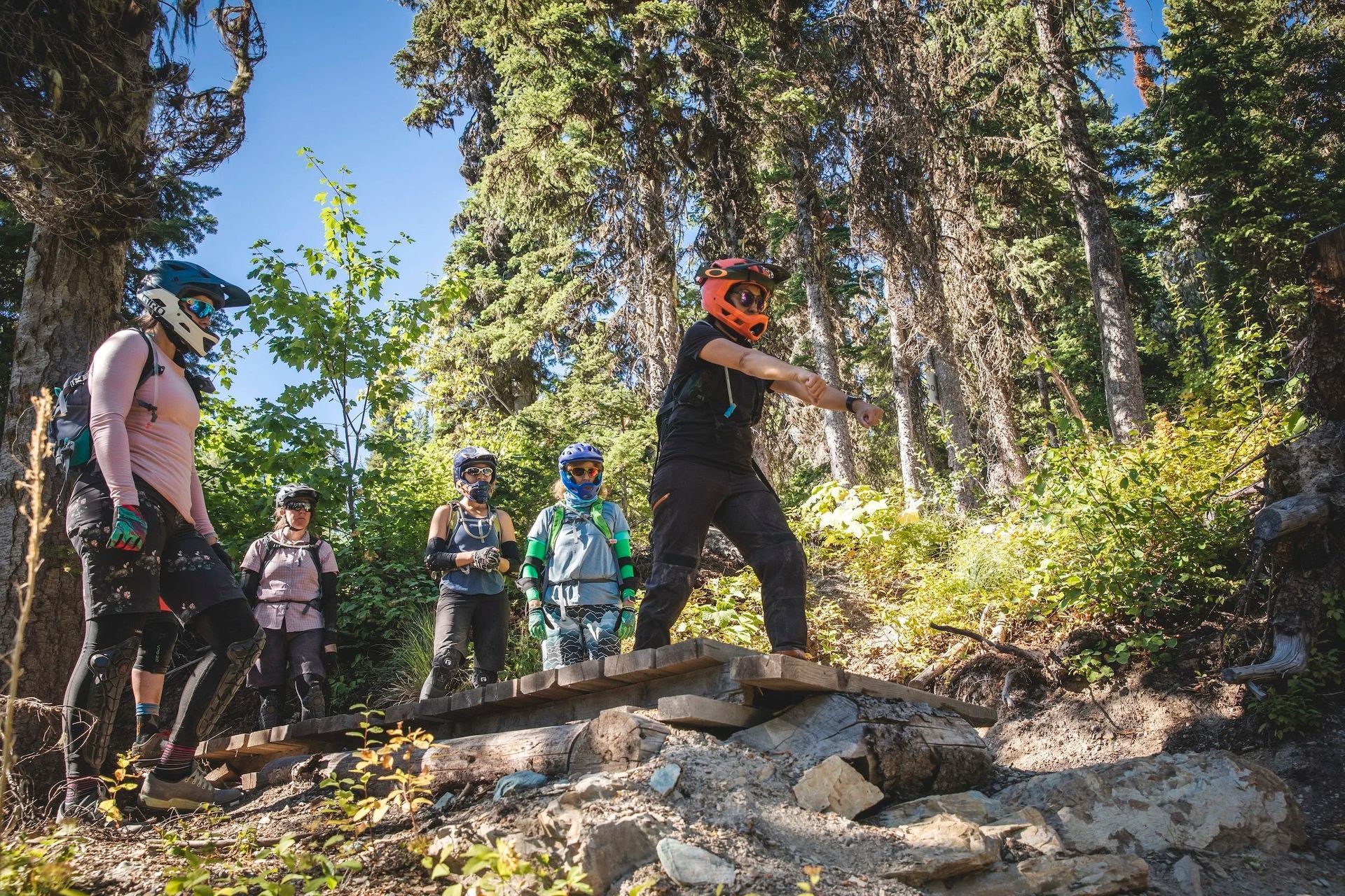 Mountain bikers on a trail, crossing a wooden bridge in a sunny forest.
