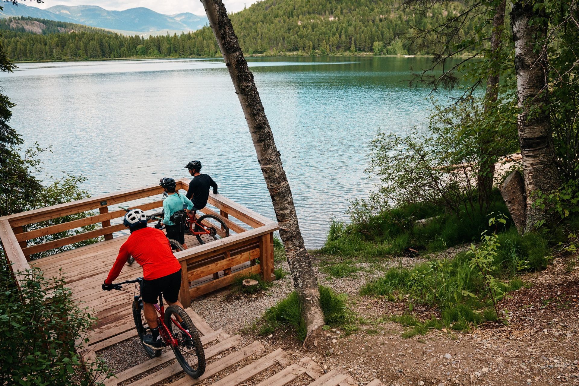 Cyclists ride bikes toward a lake on a wooden platform. Green trees and a calm body of water in the background.