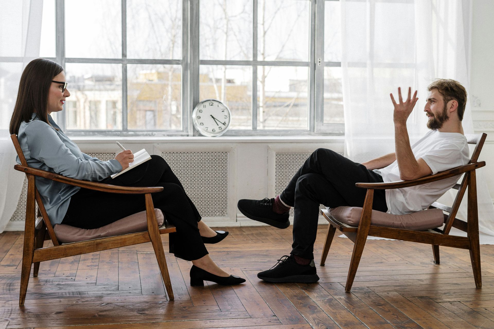 Woman therapist in glasses taking notes, talking with a man seated in front of a window.